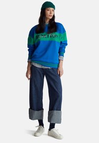 Felpa blu e verde con polsini a righe; presenta il logo "BENETTON". Pantaloni larghi in denim con orli risvoltati. Scarpe da ginnastica bianche.