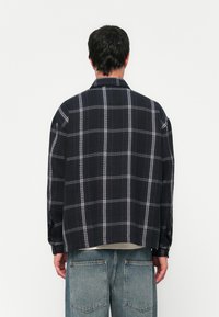 Hombre vestido con una camisa de cuadros azul marino oscuro y jeans azul claros, de pie de espaldas contra un fondo blanco liso.