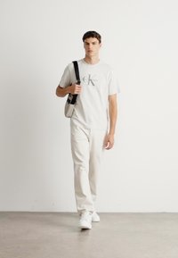 Ung mand går indendørs iført en hvid Calvin Klein t-shirt, lysebeige bukser, hvide sneakers og bærer en skuldertaske.