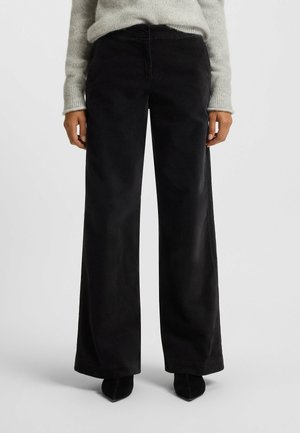 Personne portant un pantalon noir à jambes larges, un pull en maille gris et des chaussures noires pointues, debout devant un fond clair uni.