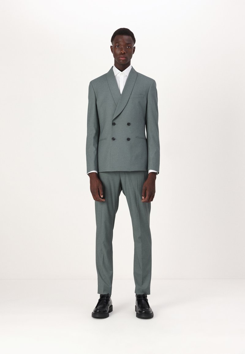 Completo doppiopetto in tessuto verde chiaro testurizzato, vestibilità tailored, con bottoni neri, abbinato a pantaloni coordinati e scarpe nere.