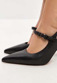 Scarpe nere in pelle con tacco alto e punta affilata, dotate di una cinghia alla caviglia e un design a intaglio sui lati. Texture liscia.
