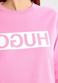 Sudadera rosa con cuello redondo, que presenta un atrevido texto blanco "HUGO" en la parte frontal. Textura suave, corte casual.