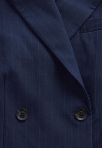 Tengerészkék tűszegélyes dupla soros blézer sima anyagból, nagy fekete gombokkal és egyetlen mellzsebbel.