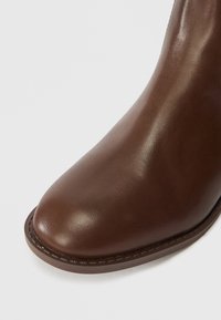 Botim de tornozelo em couro castanho com bico arredondado, textura suave e detalhe de costura visível ao longo da sola.