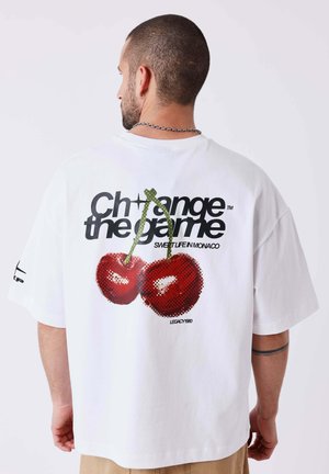 Uomo con capelli corti che indossa una maglietta bianca con grandi ciliegie e la scritta "Change the game" sulla schiena, di spalle.