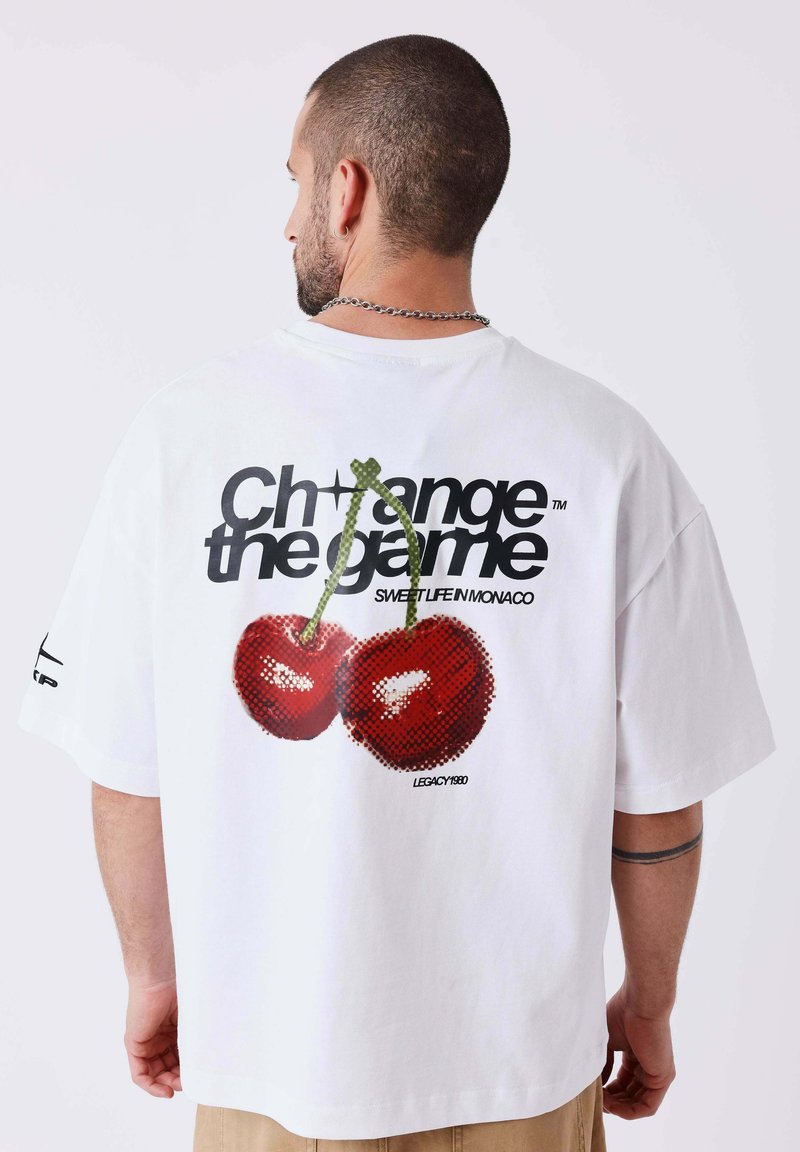 Homme aux cheveux courts portant un t-shirt blanc avec de grosses cerises et le texte « Change the game » dans le dos, vu de dos.