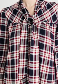 Blusa in cotone con motivo a quadri nei colori blu navy, rosso, grigio e bianco. Presenta un colletto a fiocco e un dettaglio di tessuto arricciato sul petto.