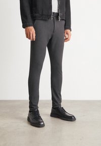 Man in een slim-fit donkergrijze broek, zwarte riem, zwarte leren schoenen en een zwart jasje, staande op een gladde betonvloer.