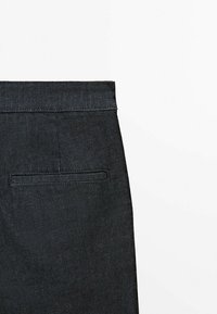 Pantalon en denim noir avec une ceinture plate, une poche arrière unique et des détails de couture subtils. Le tissu a une texture lisse.