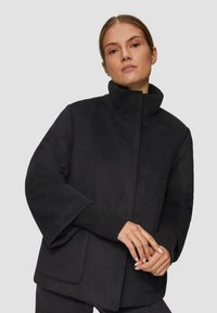 Czarny płaszcz z mieszanki wełny z wysokim kołnierzem, szerokimi rękawami, ściągaczami na mankietach i bocznymi kieszeniami. Gładka struktura i minimalne detale szwów.