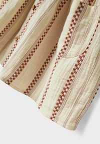 Krämfärgad tyg med vertikala bruna rutiga och prickiga ränder, synliga knappar och en fastsydd ficka på texturerat material.