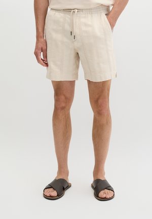 Mężczyzna w beżowych, teksturowanych spodenkach ze ściągaczem i czarnych sandałach na wsuwkę, stojący z rękami w kieszeniach na jednolitym tle.