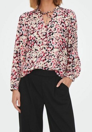 Kobieta nosząca bluzkę z długim rękawem z abstrakcyjnym kwiatowym wzorem w kolorach czerwonym, różowym, czarnym i białym, w połączeniu z luźnymi czarnymi spodniami.