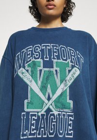 Blå överdimensionerad sweatshirt med texten "WESTFORT LEAGUE" och ett stort grönt "W," med korslagda basebollträn i sliten tryck.