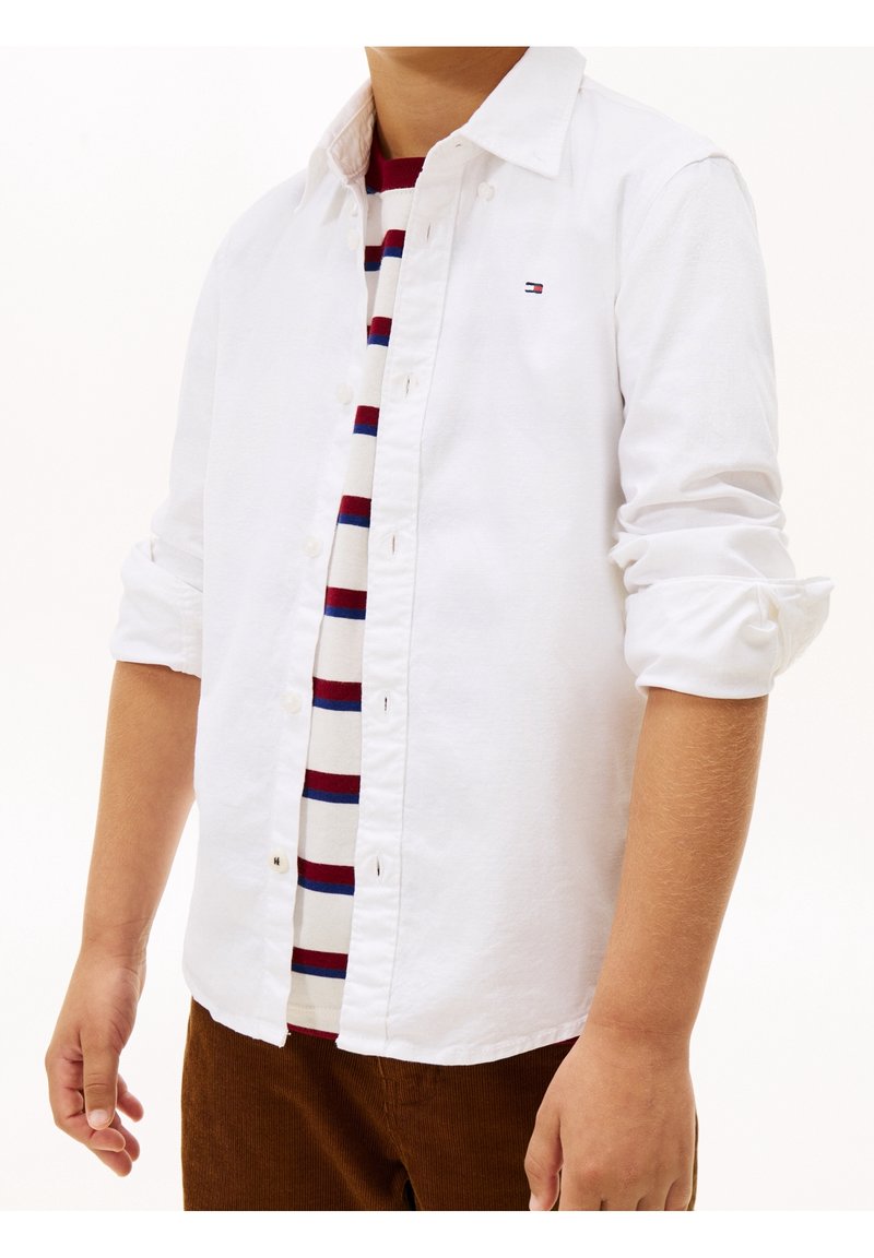 Personne portant une chemise blanche boutonnée sur un t-shirt à rayures rouges, bleues et blanches, avec les manches retroussées et un pantalon marron visible.