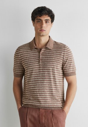 Ung mand med krøllet mørkt hår iført en brun og beige stribet kortærmet polo-shirt og brune bukser, står med hænderne i lommerne.