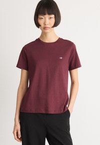 Woman with short bob haircut wearing a plain burgundy short-sleeve t-shirt and black pants, standing against a white background.