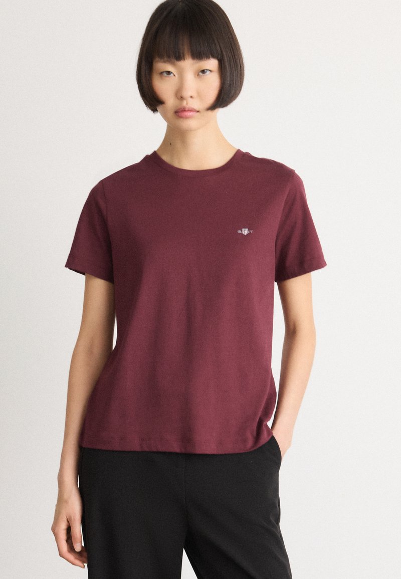 Woman with short bob haircut wearing a plain burgundy short-sleeve t-shirt and black pants, standing against a white background.