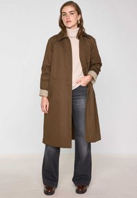 Cappotto lungo marrone con colletto, chiusura con bottoni e tasche laterali. Sotto, un dolcevita beige chiaro e jeans grigi a zampa.