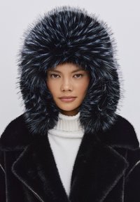 Sort faux fur hætte med lang, fluffy struktur, indrammet af en hvid sweater med høj krave og en slank sort frakke.
