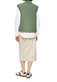 Gilet matelassé vert avec col montant porté sur une chemise blanche, associé à une jupe beige longueur genoux et des baskets décontractées avec des accents noirs.
