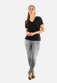 Maglietta nera con scollo a V, maniche corte; abbinata a jeans skinny grigio chiaro e tacchi alti neri. Modella in piedi con le mani incrociate, sfondo neutro.