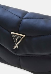 Borsa nera in ecopelle con texture trapuntata, caratterizzata da un logo GUESS triangolare oro e dettagli hardware decorativi.