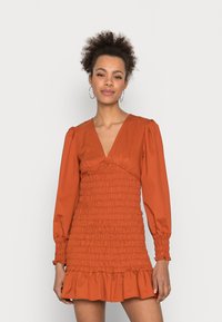 Vestido de cor ferrugem com mangas bufantes longas, decote profundo em V e textura franzida, terminando em uma bainha com volantes à altura da coxa.