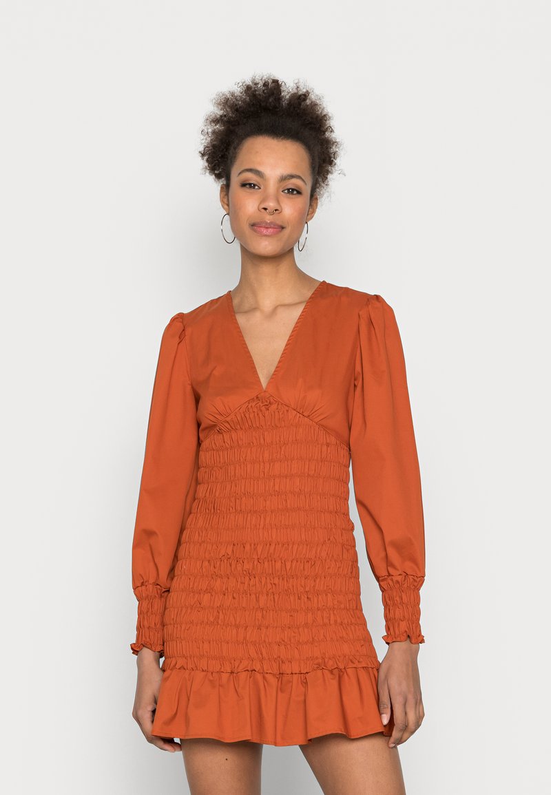 Vestido de cor ferrugem com mangas bufantes longas, decote profundo em V e textura franzida, terminando em uma bainha com volantes à altura da coxa.