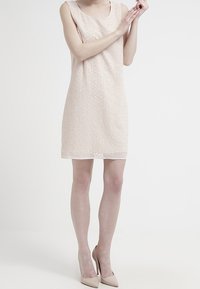 Robe sans manches rose clair avec motif géométrique texturé, décolleté rond et coupe trapèze, associée à des talons nus. Design minimaliste.