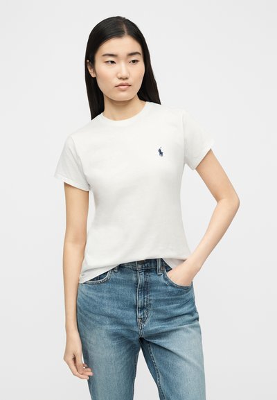 Νέα γυναίκα με μακριά σκούρα μαλλιά φοράει λευκό T-shirt με κοντά μανίκια και λογότυπο και μπλε τζιν, στέκεται με το ένα χέρι στην τσέπη μπροστά σε απλό φόντο.