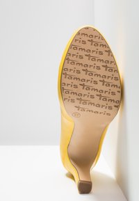 Chaussure jaune à talons hauts montrant la semelle avec un motif répétitif du logo "Tamaris" et la pointure 37 embossée près du talon.
