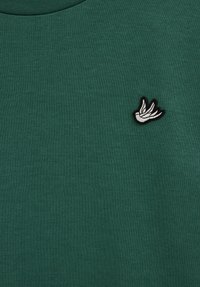 Pull vert foncé avec une texture côtelée, présentant un petit logo brodé d'un oiseau noir et blanc sur la partie gauche de la poitrine.
