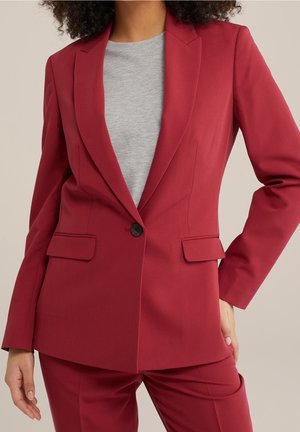 Vrouw die een op maat gemaakte rode blazer draagt over een lichtgrijs overhemd, met bijpassende rode broek, staand met één hand op haar heup.