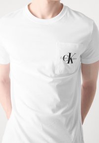 Muž v přiléhavém bílém tričku s krátkým rukávem a malou kapsičkou na levé straně hrudníku, na které je logo "Calvin Klein Jeans".