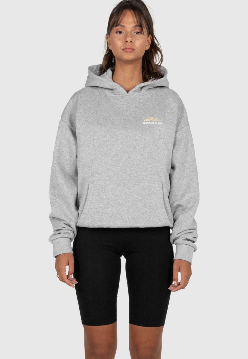 Sweat-shirt à capuche gris avec une poche avant, arborant un petit logo de montagne en doré, associé à un short de vélo noir. Texture lisse.
