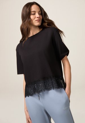 Zwarte crop top met korte mouwen, kant-detail aan de onderrand en geborduurde tekst. Gecombineerd met lichtblauwe broek met een tekstdetail.