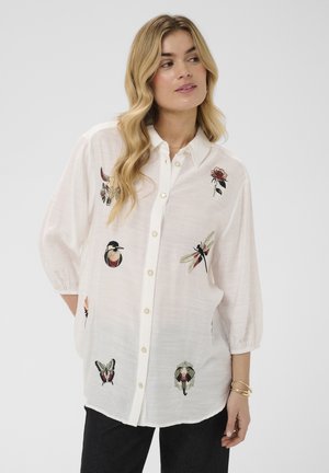 Kvinde iført en hvid skjorte med knapper med broderede insekter, fugle og blomster, kombineret med sorte bukser og guldbånd.
