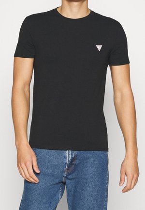 Homme portant un t-shirt noir uni à manches courtes avec un petit logo triangulaire blanc et rouge sur le côté gauche de la poitrine, associé à un jean en denim bleu.