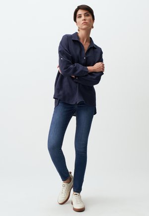 Camicia in lino blu navy con colletto e dettagli di bottoni, abbinata a jeans scuri slim fit e sneakers bianche con accenti marroni.