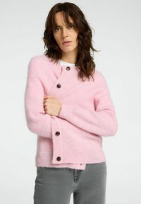 Cardigan en tricot rose avec une texture douce, encolure ronde et deux boutons sombres à l'avant. Porté sur une chemise blanche, avec des manches longues.