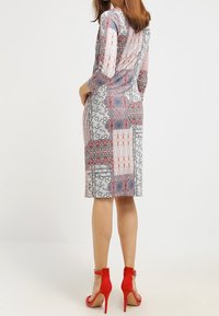 Dvojbarevné vzorované šaty s tříčtvrtečními rukávy a délku do kolen; doplněné červenými lodičkami na vysokém podpatku.