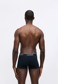 Black boxer shorts with a white Lacoste logo on the waistband, made from a smooth fabric. Model shown from the back, with no additional details.