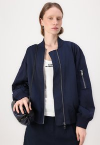 Ναυτική μπόμπερ ζακέτα με φερμουάρ, ριμπ μανσέτες και πλευρική τσέπη. Φοριέται πάνω από ένα λευκό μπλουζάκι, κρατώντας μια σκούρα τσάντα.