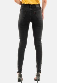 Pantalones vaqueros skinny negros de mezclilla, con una cintura alta, dos bolsillos traseros y una textura ligeramente desgastada, combinados con tacones altos negros.