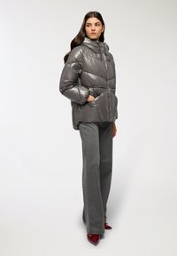 Giacca a vento grigia con finitura lucida, dotata di cappuccio, chiusura con zip e tasche laterali, abbinata a pantaloni grigi a righe e tacchi rossi.