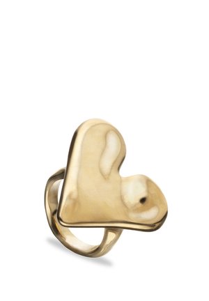 Anillo tono dorado con parte superior ancha y asimétrica en forma de corazón y superficie lisa y reflectante sobre un fondo blanco.