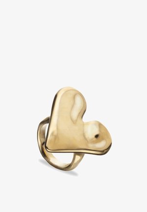 Anillo tono dorado con parte superior ancha y asimétrica en forma de corazón y superficie lisa y reflectante sobre un fondo blanco.