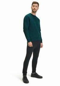 Felpa verde scuro a girocollo con maniche a costine, abbinata a pantaloni neri slim-fit e sneakers nere con lacci, su uno sfondo bianco.